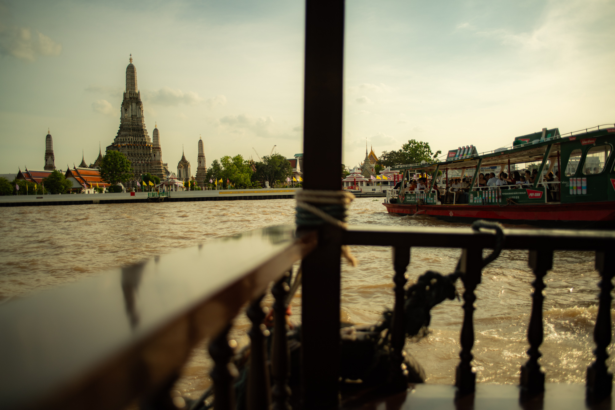 Vista desde un barco en un río con el templo de Wat Arun al fondo y un transbordador de pasajeros pasando en primer plano, bajo un cielo parcialmente nublado.
