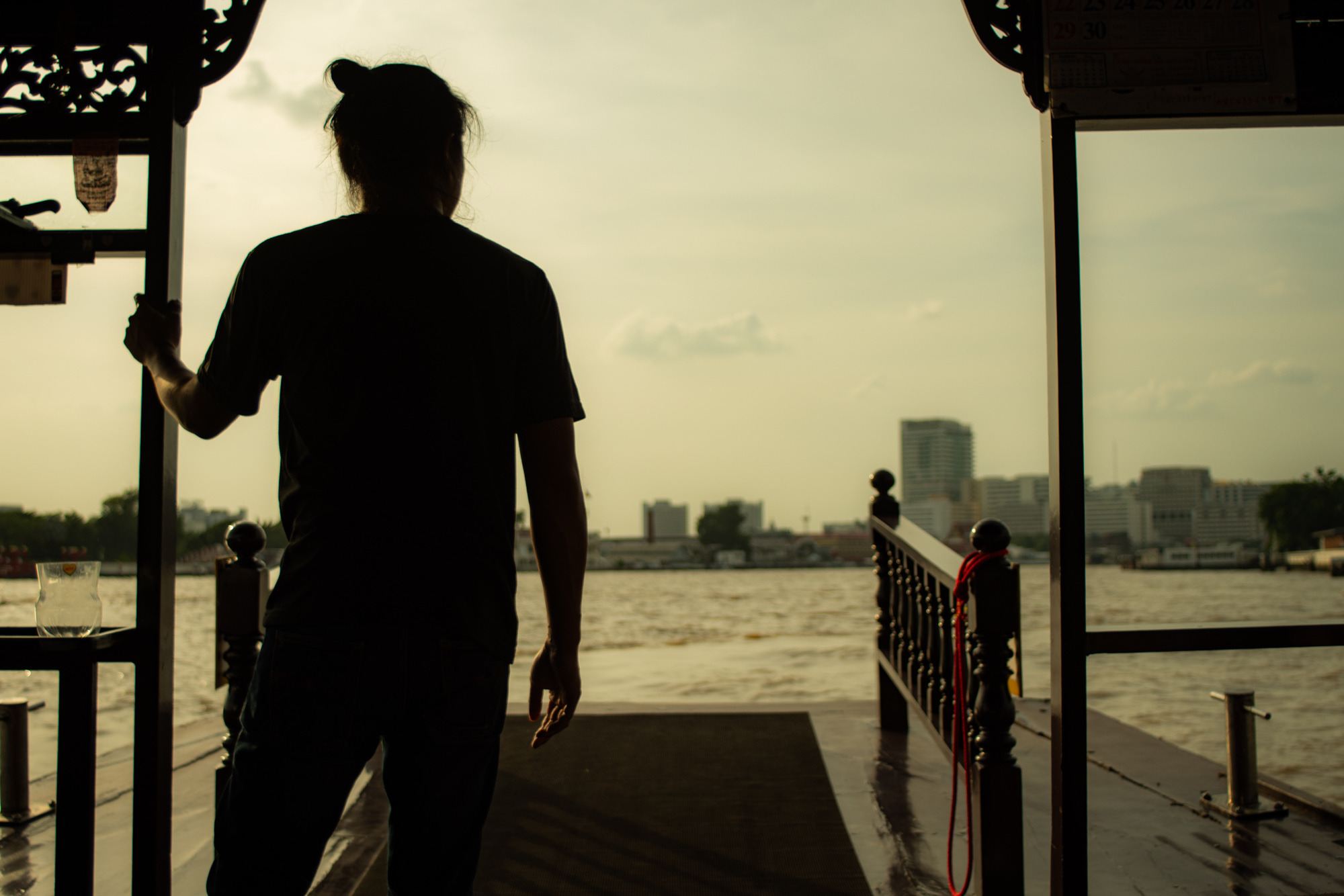 Silueta de una persona de pie en el borde de un barco mirando hacia un río con un horizonte de la ciudad en el fondo durante la puesta de sol.