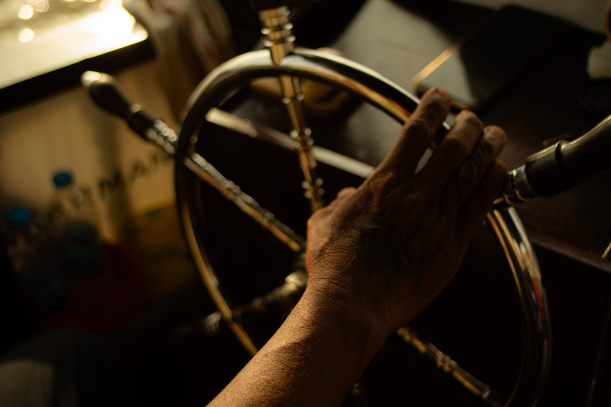 La mano de una persona agarra la rueda metálica de un barco o una embarcación, con la luz del sol entrando por una ventana cercana.