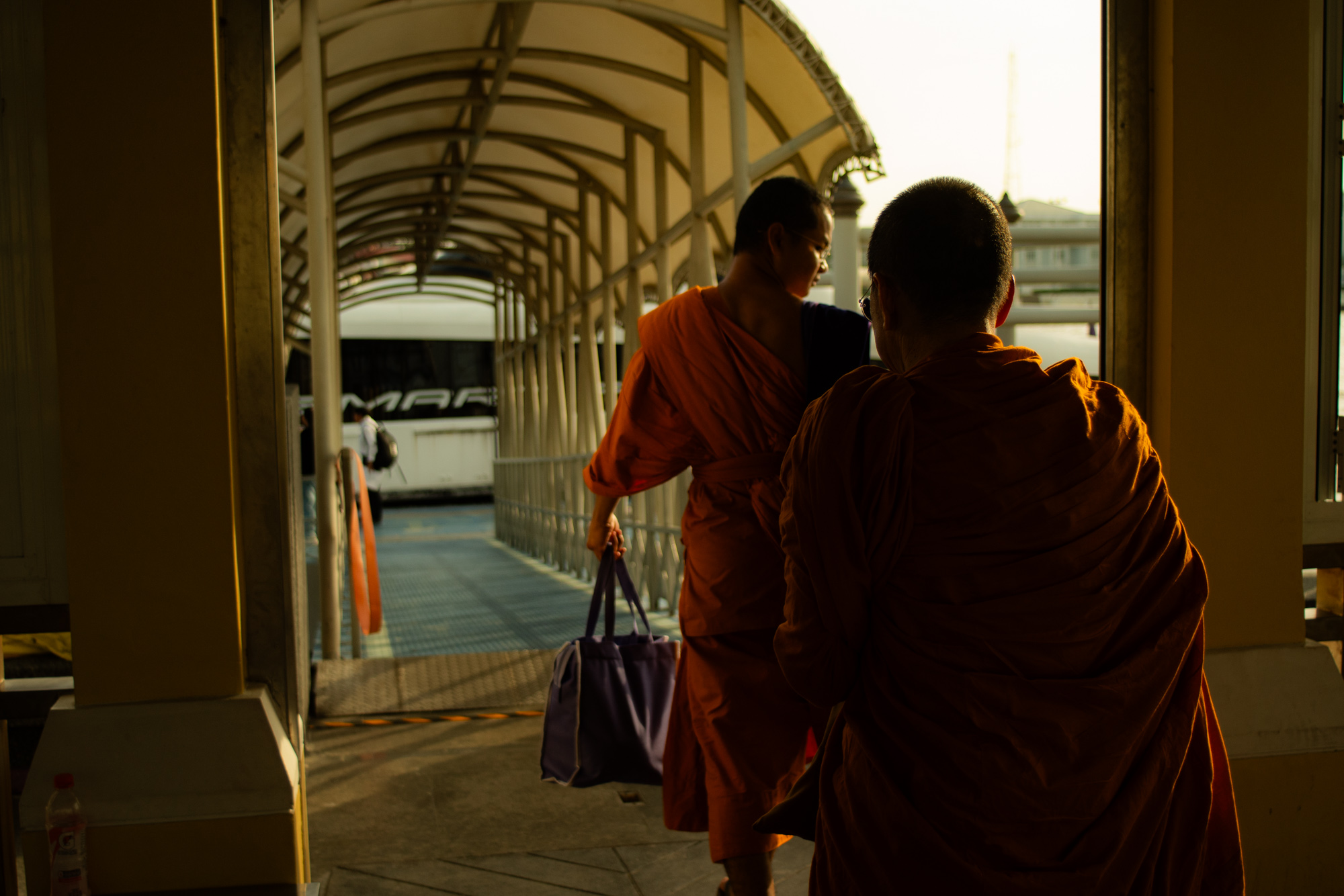 Dos personas vestidas con túnicas naranjas caminan por un pasillo cubierto hacia un autobús; una de ellas lleva una bolsa morada. La luz del sol proyecta largas sombras.