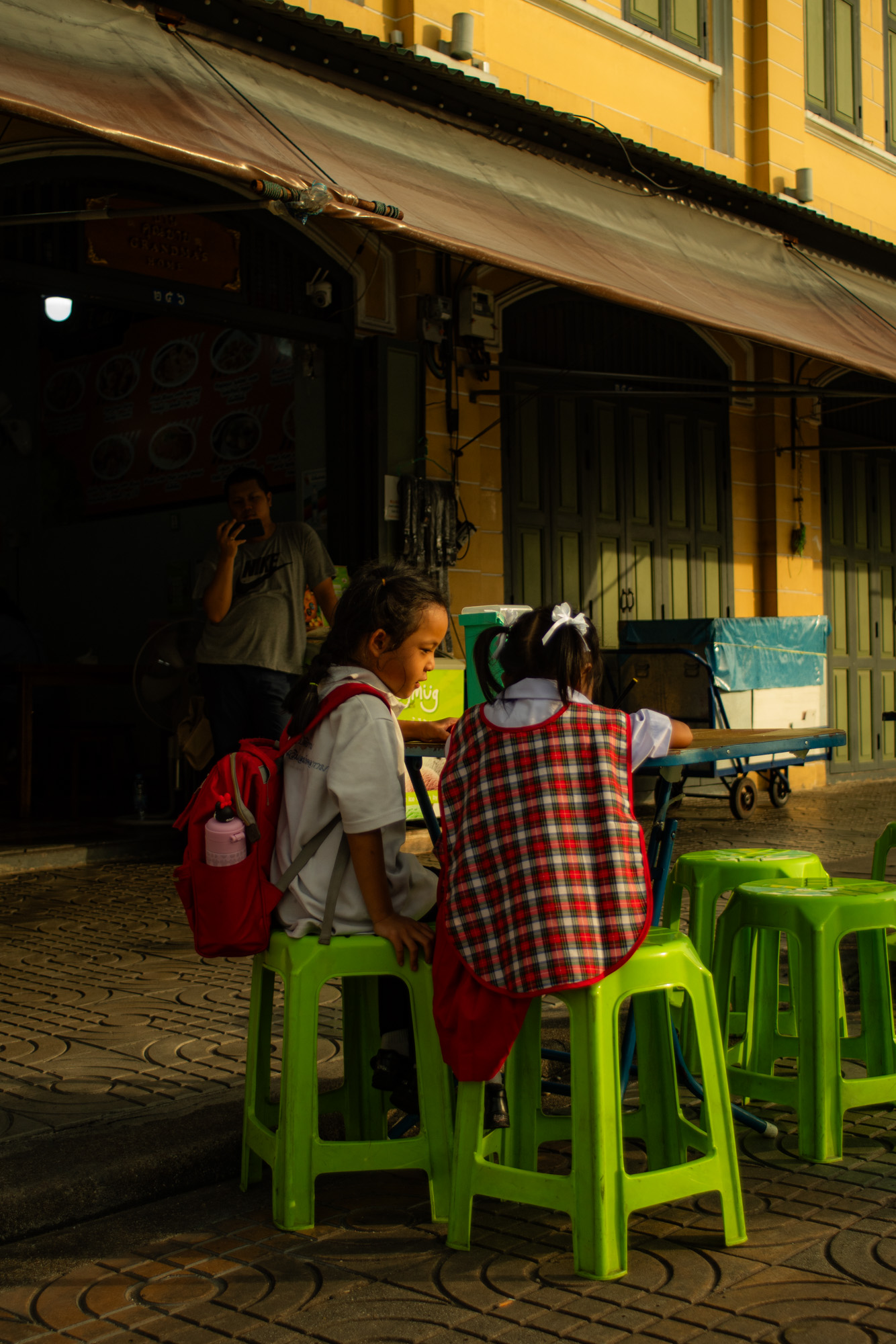 Dos niños con uniforme escolar se sientan en taburetes de plástico verde en un puesto de comida al aire libre; uno lleva una mochila roja, y una persona permanece de pie al fondo, cerca de la entrada de un edificio.