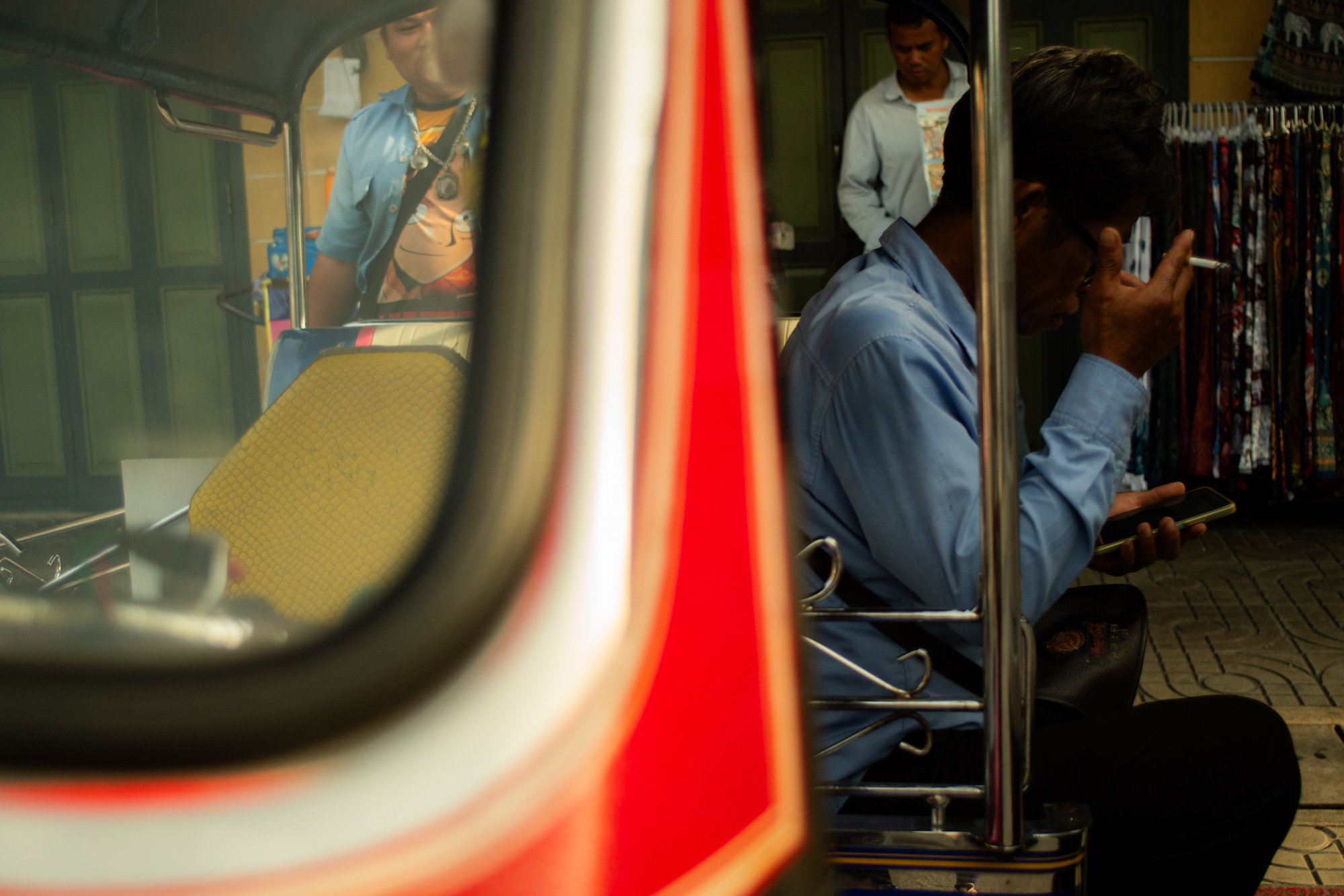 Un hombre con camisa azul va sentado en un rickshaw, con un cigarrillo en la mano y consultando su teléfono; otra persona permanece de pie al fondo, cerca de tiendas de colores.