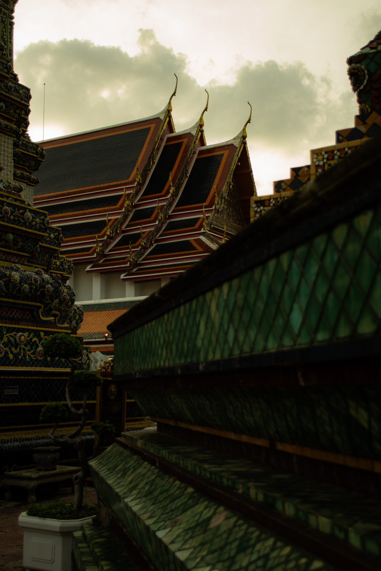 Techos de templos tradicionales tailandeses con detalles ornamentales, vistos al atardecer con tejas verdes decorativas en primer plano y un cielo nublado en lo alto.