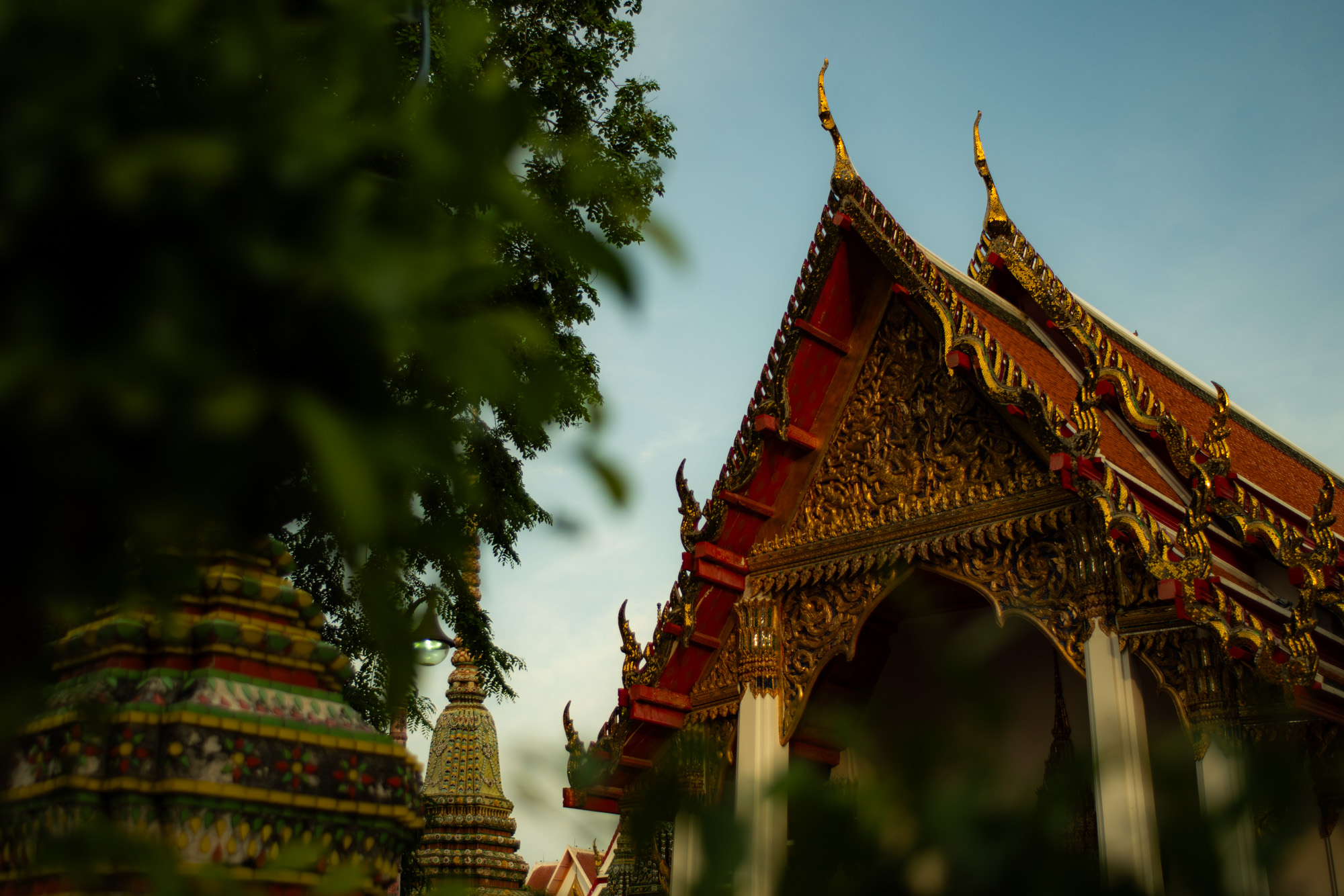 Templo tailandés ornamentado con intrincados detalles dorados y tejado puntiagudo, parcialmente oculto por la vegetación, con chedis visibles al fondo bajo un cielo despejado.