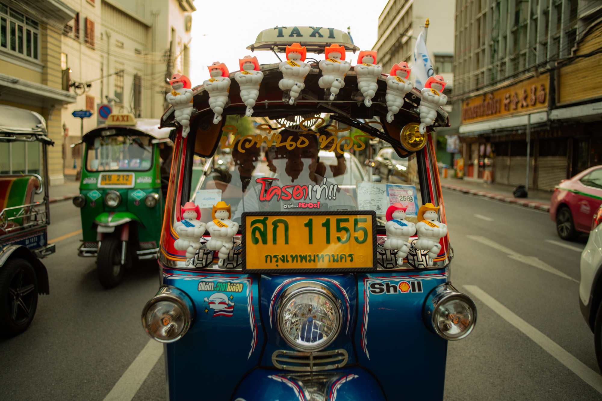 Un tuk-tuk azul decorado con muñecas de Hello Kitty aparcado en una calle de Tailandia, con otros tuk-tuks y edificios al fondo.