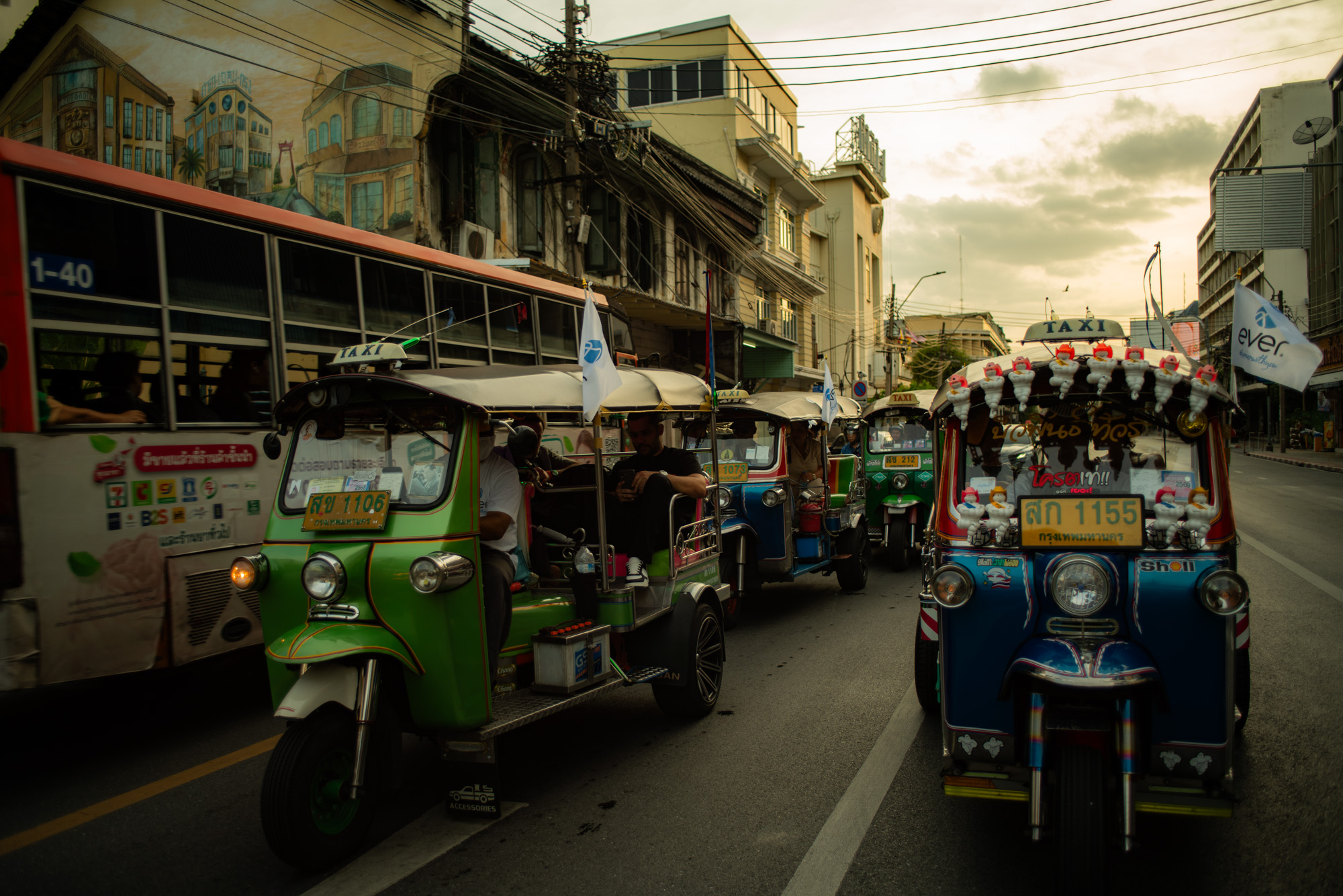 Tuk-tuks y un autobús urbano recorren una concurrida calle bordeada de edificios y cables aéreos bajo un cielo nublado al atardecer.