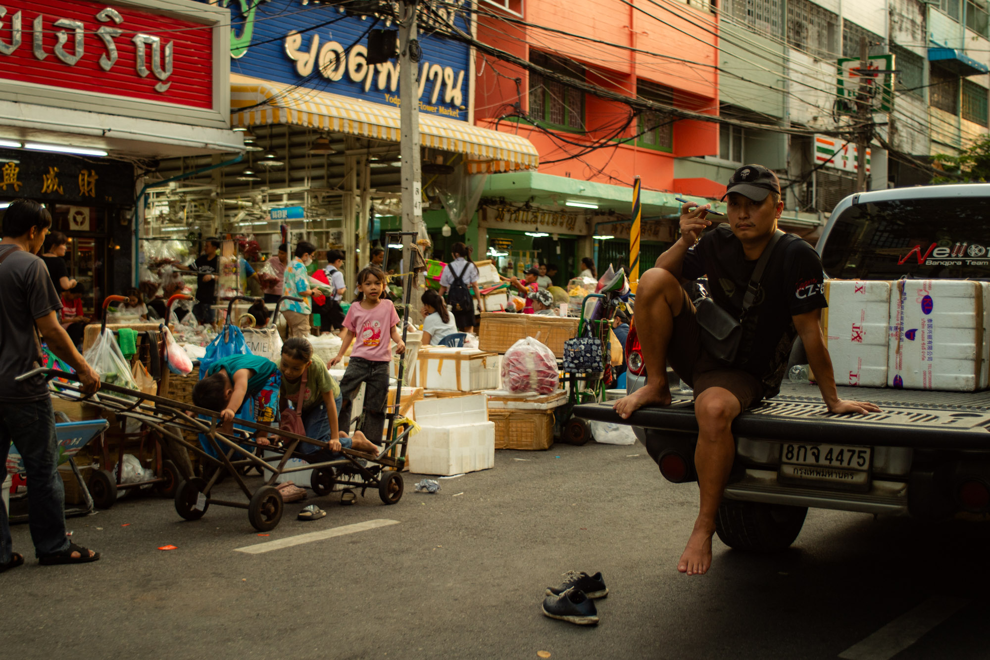Un hombre se sienta descalzo en la parte trasera de un camión mientras la gente mueve carros y mercancías en una escena callejera de un ajetreado mercado al aire libre.