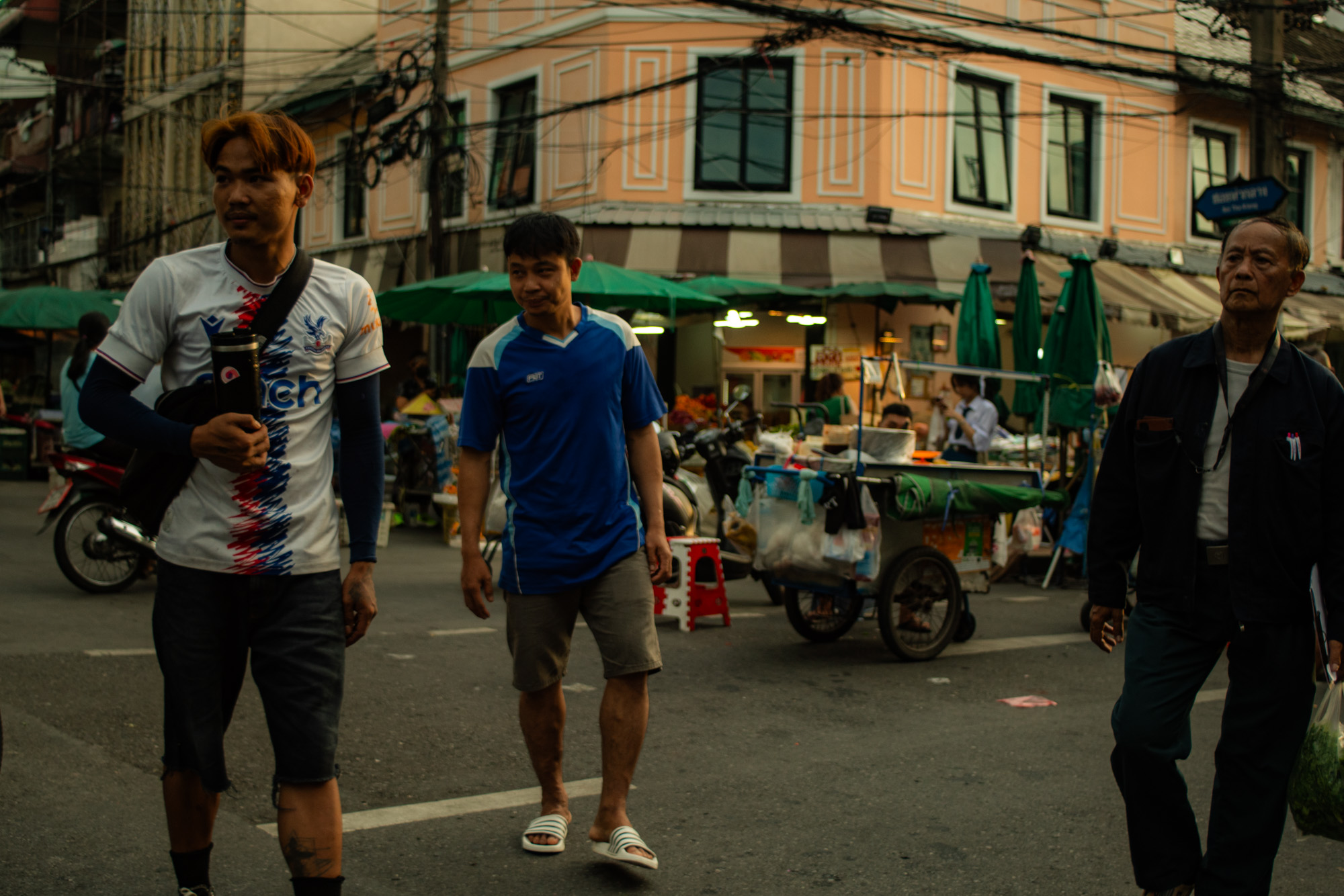 Tres hombres caminan por una calle con puestos de mercado y un edificio al fondo; uno de ellos lleva una camiseta deportiva, otro viste de azul y el tercero lleva ropa oscura.