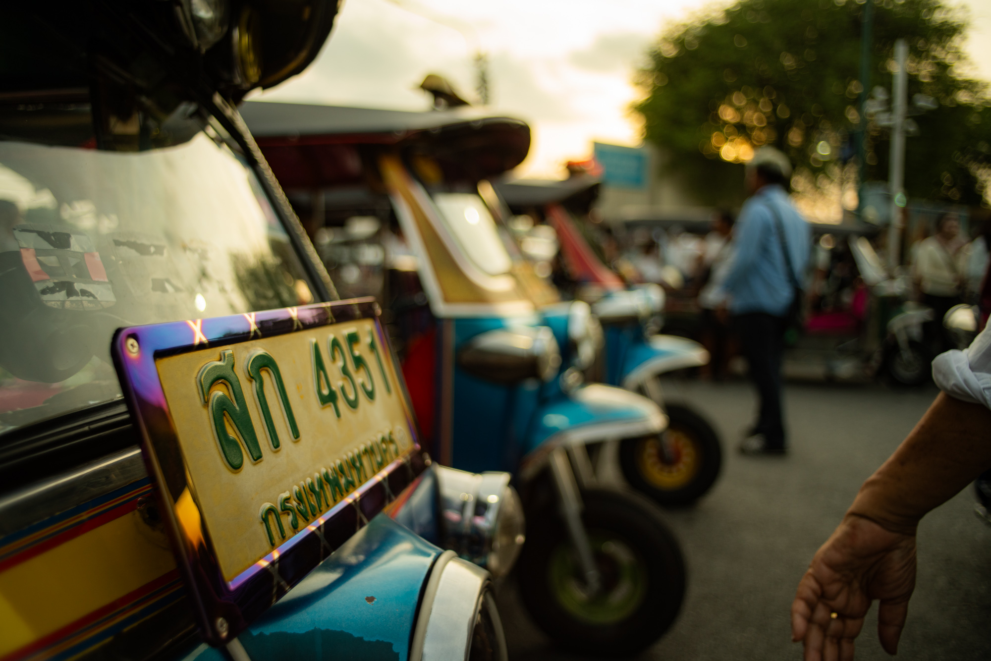 Primer plano de la matrícula de un tuk-tuk enfocada, con varios otros tuk-tuks y gente al fondo en una calle al atardecer.