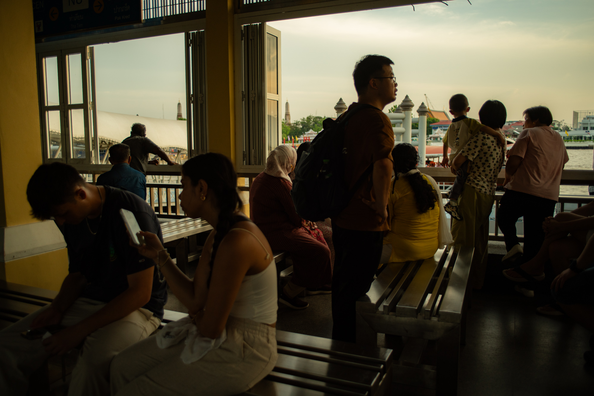 La gente se sienta y se para en los bancos de una terminal de transbordadores, algunos mirando sus teléfonos, otros observando el río y el paisaje urbano a través de las puertas abiertas al atardecer.