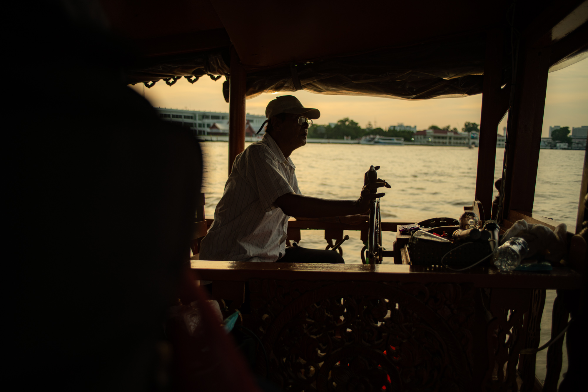 Una persona con sombrero se sienta al timón de una barca en un río, silueteada contra el agua y los edificios lejanos al atardecer.