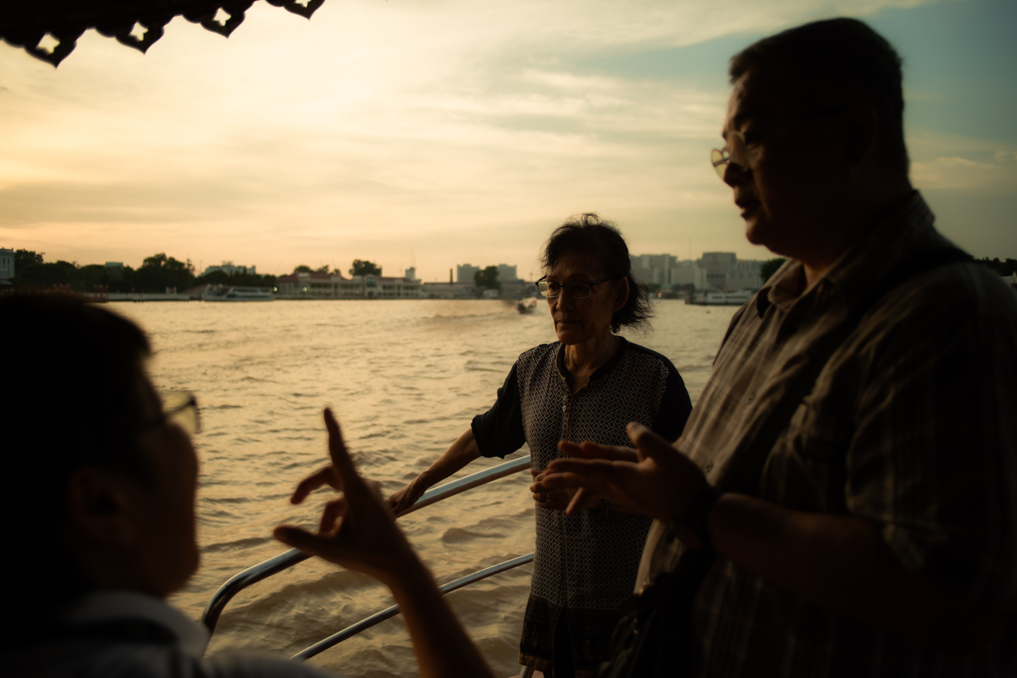 Tres personas conversan en un barco al atardecer, con un río y el perfil de la ciudad visibles al fondo.