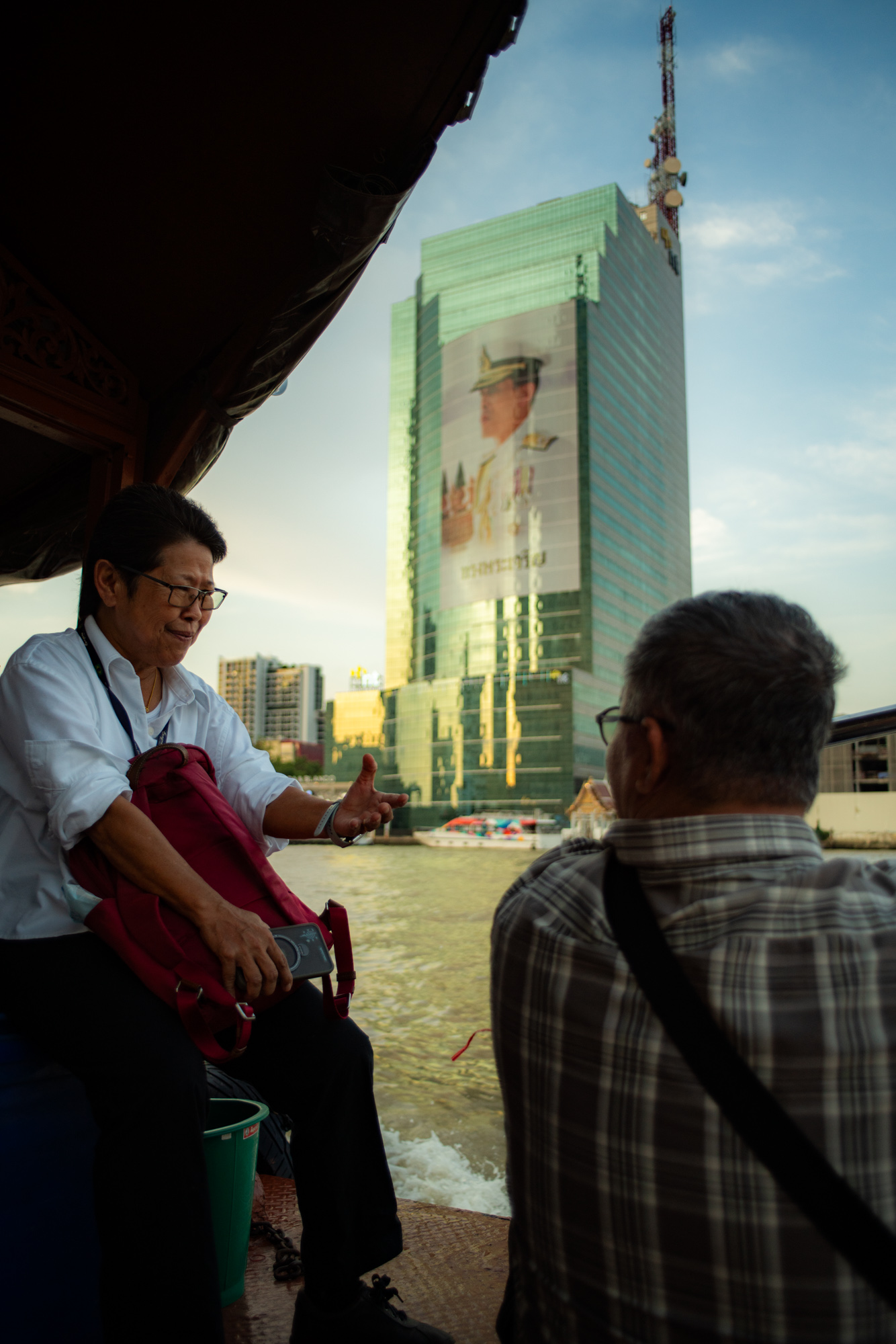 Dos personas sentadas en una barca con un río al fondo; al otro lado del agua se ve un moderno edificio de cristal con un gran anuncio.