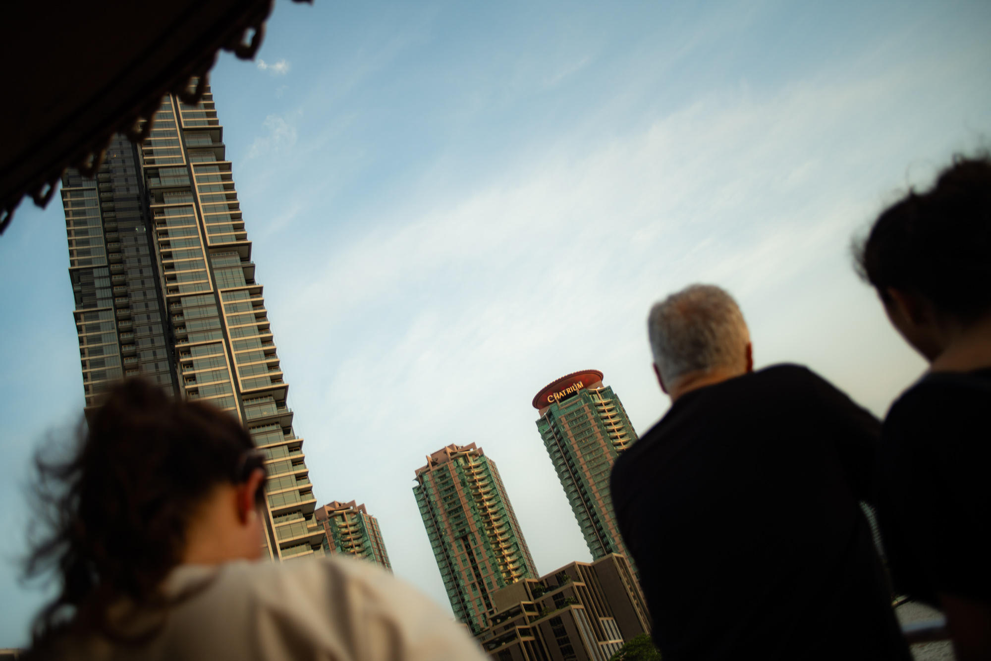 Tres personas se sitúan en primer plano frente a altos edificios modernos, entre ellos uno con un cartel de Chatrium, bajo un cielo despejado a la luz del día.