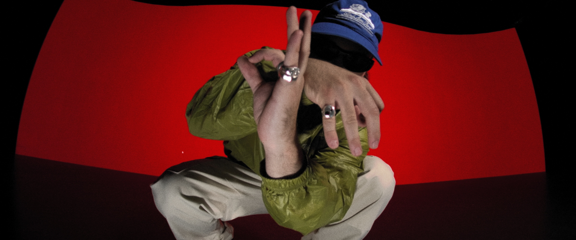 Una persona con gorra azul, gafas de sol, chaqueta verde y pantalones beige se agacha con las manos levantadas ante un fondo rojo.