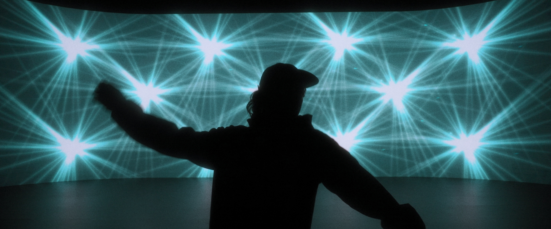 Una persona en silueta está de pie con un brazo levantado delante de una pantalla que muestra un patrón de estrellas azules brillantes.